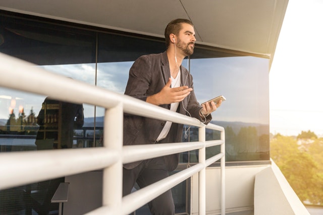 Muž v hnedom saku stojí na balkóne opretí pri zábradlí a telefonuje cez handsfree